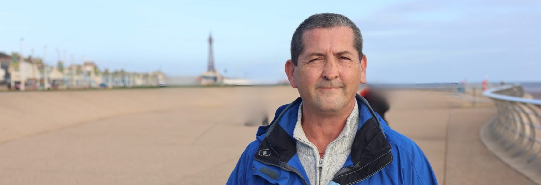 A man named Ian wearing a blue coat standing by the pier near the beach. Sky is blue and it's a pleasant day.