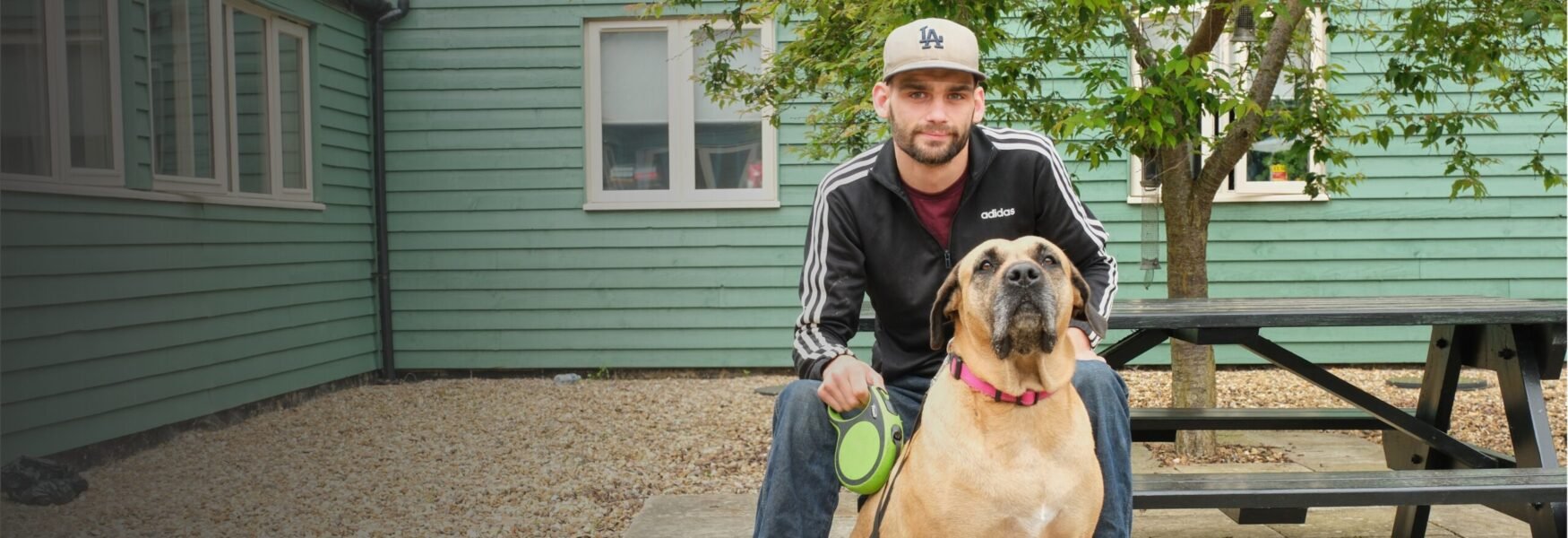 A young man sits on a bench outside a green building with a large tan dog on a leash beside him.